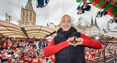 Gebärdensprachdolmetscher Michael Zymelka steht auf der Bühne mit dem Rücken zum Publikum und lacht in die Kamera. Es ist Weiberfastnacht auf dem Alter Markt in Köln.