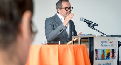Markus Bachmann am Rednerpult mit Banner Wir gestalten Barrierefreiheit und dem Logo der Agentur Barrierefrei NRW