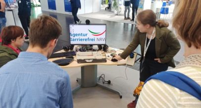 Ein Schreibtisch mit einem Bildschirm. Auf dem Bildschirm das Logo der Agentur Barrierefrei NRW. Auf dem Schreibtisch liegen viele adaptive Hilfsmittel. Daneben steht Frau Laumann und zeigt drei von hinten aufgenommenen Personen die Hilfsmittel.