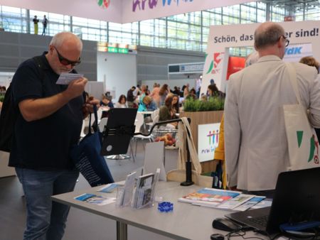 Infotisch der Agentur Barrierefrei NRW bei der REHACARE. Ein Mann steht davon und schaut sich genau eine Postkarte mit Tipps zur Barrierefreiheit an.