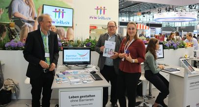 Messeauftritt, Schriftzug NRW inklusiv und Logo der Agentur Barrierefrei NRW auf einem Bildschirm, drei Menschen an einem Tisch, Tisch mit Broschüren