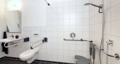 Barrierefreies Bad mit Hilfsmitteln wie beidseitigen Haltegriffen am WC, eine befahrbaren Dusche mit klappbaren Sitzhocker, unterfahrbarem Waschtisch