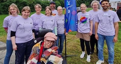 Gruppenbild mit insgesamt 10 Personen, eine davon im Rollstuhl, die in einheitlichen T-Shirts neben dem Messestand um die Fahne der KSL NRW stehen und in die Kamera lächeln.