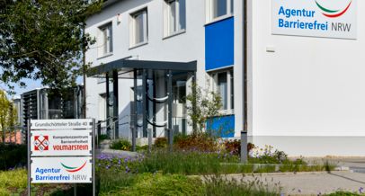 Außenansicht des Gebäudes mit den Wegweisern KBV und Agentur unter dem Schild Grundschötteler Straße 40 im blühenden grünen Vorgarten des blau-weißen Gebäudes mit einem großen Banner der Agentur an der seitlichen Hauswand