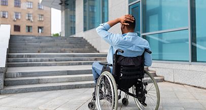 Rollstuhlfahrer steht fragend vor dem Teppenaufgang eines Gebäudes.