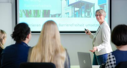 Ein Seminarraum mit Blick zur Leinwand, davor Mitarbeiterin Lydia Weyandt. Sie steht als Dozentin vor der Leinwand und blickt die Teilnehmenden mit einem Lächeln an. Diese sieht man nur von hinten.