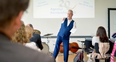 Moderator Rainer Schmidt im Hintergrund Schriftzug Festveranstaltung 20 Jahre Agentur Barrierefrei NRW