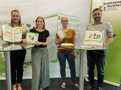 Foto: Das Projektteam steht an zwei Stehttischen. Im Hintergrund ist eine Stellwand mit dem Logo von der Ärztekammer Nordrhein. Das Projektteam hält die Materialien in Leichter Sprache in den Händen und in die Kamera.