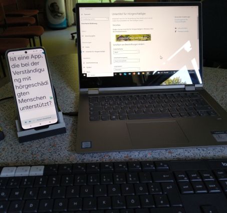 An einem PC Arbeitsplatz läuft eine KI basierte App auf dem Smartphone zur automatischen Transkription. Das Smartphone seht in einem Stativ.