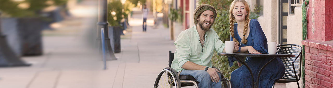 Ein junger Mann im Rollstuhl und eine junge Frau sitzen an einem Bistrotisch in einem Straßencafe und lacenh in die Kamera